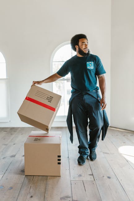 A male removal worker with dark curly hair and a beard, dressed in a navy blue t-shirt with a light blue geometric logo on the chest, is inside a bright, minimally decorated room with white walls and 
