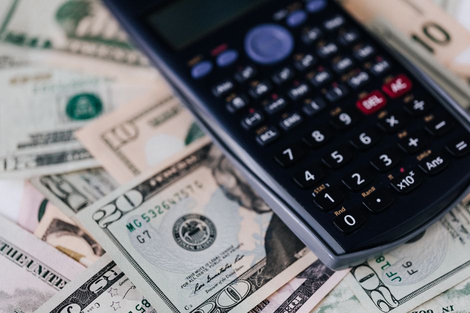 Close-up image showing a scientific calculator placed on top of scattered US dollar bills of various denominations, including twenty-dollar and ten-dollar notes. The calculator, with multiple buttons