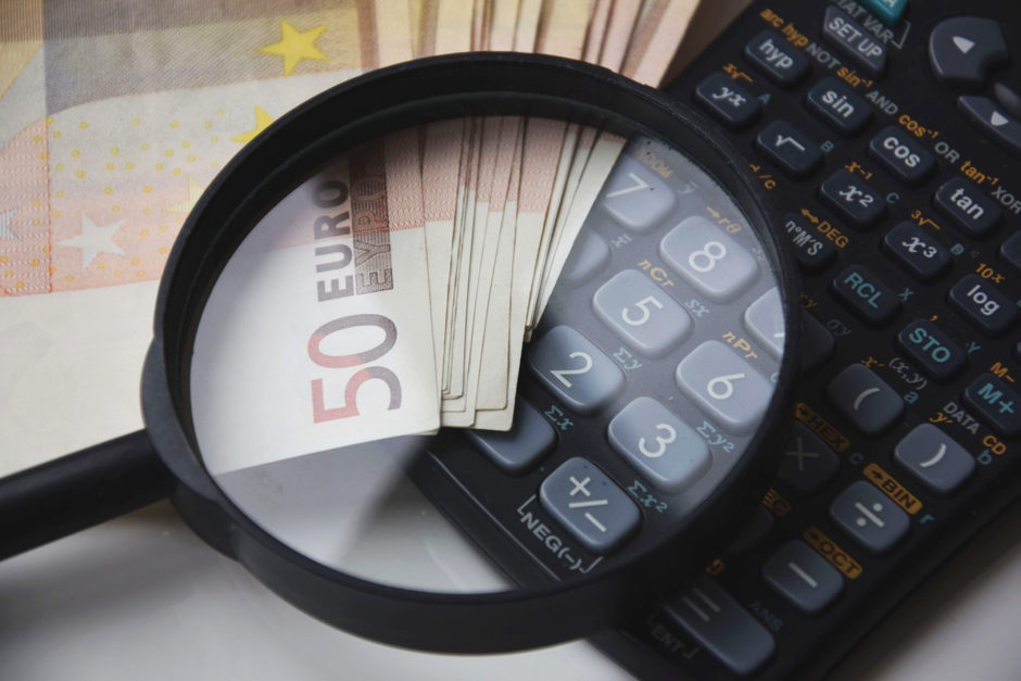 Close-up image showing a scientific calculator placed on top of scattered US dollar bills of various denominations, including twenty-dollar and ten-dollar notes. The calculator, with multiple buttons