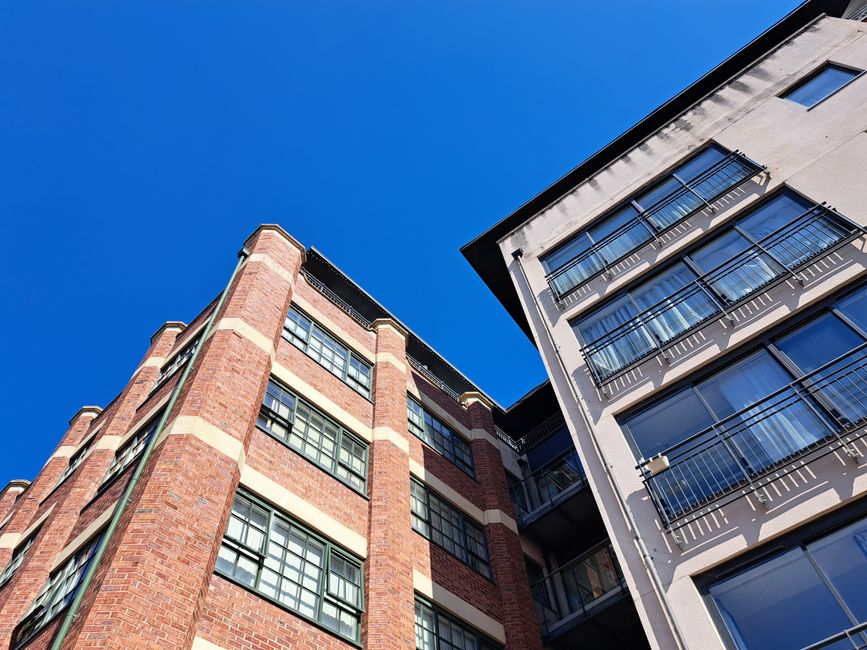 A low-angle view of two residential buildings against a clear blue sky, with the building on the left constructed from red brick featuring several large windows with dark frames, and the building on t