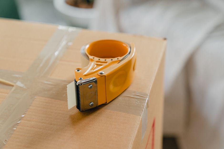 A close-up view of an orange packing tape dispenser resting on top of a cardboard moving box, taped securely with clear packing tape to prepare fragile collections for home relocation. The scene is in